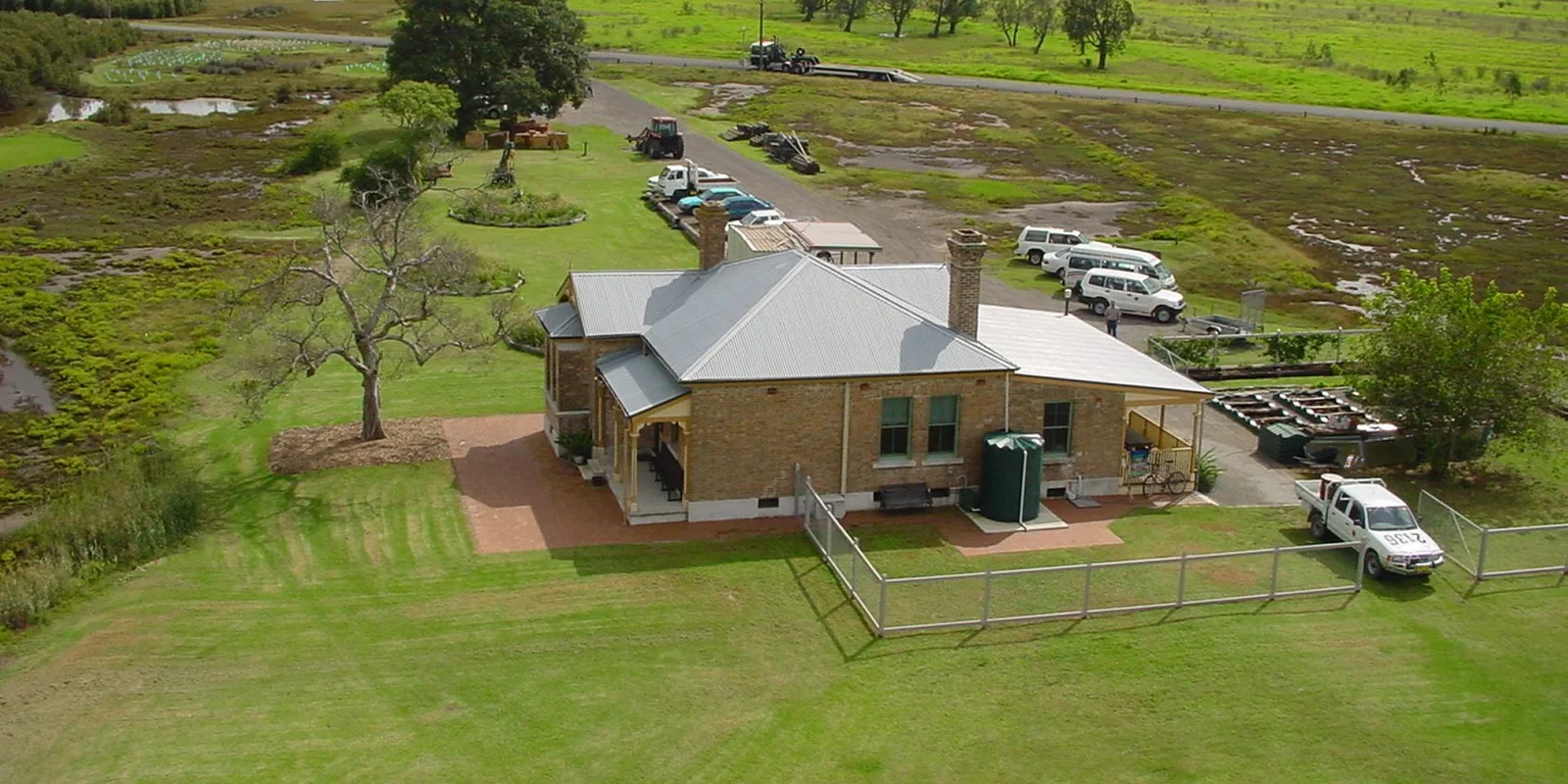 Aerial view of the Schoolmasters House and its fenced yard standing alone among wetland and paddocks