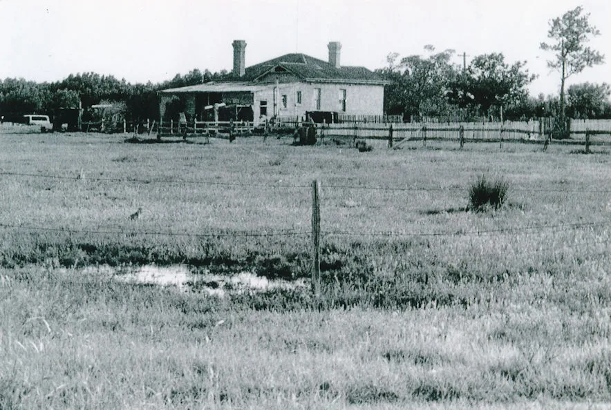 Black and white photograph of the brick Schoolmasters House behind farm fences and paddocks