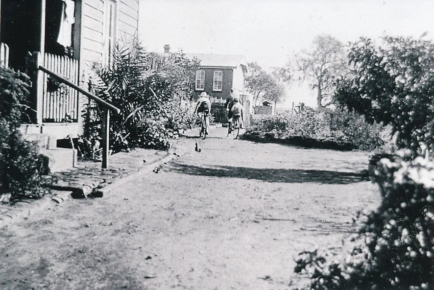 Black and white photograph of two children riding bicycles up a garden path between houses
